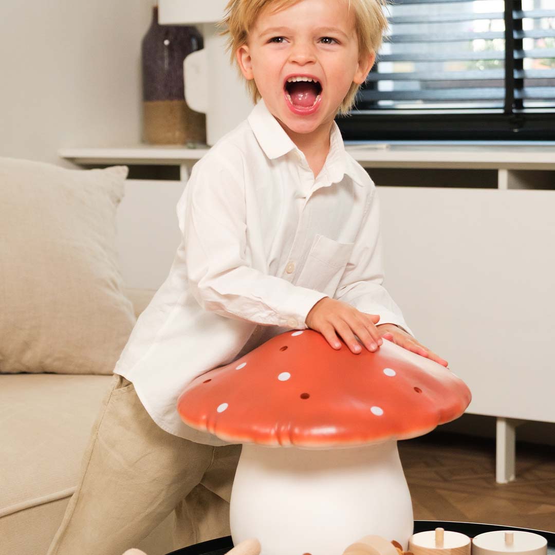 Een lachende jongen leunt op een grote Egmont Toys vliegenzwam nachtlamp met rode kap en witte stippen.
