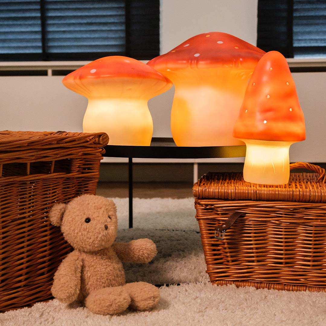 Drie Egmont Toys vliegenzwam nachtlampen in verschillende formaten, brandend op een salontafel naast rieten manden en een teddybeer.