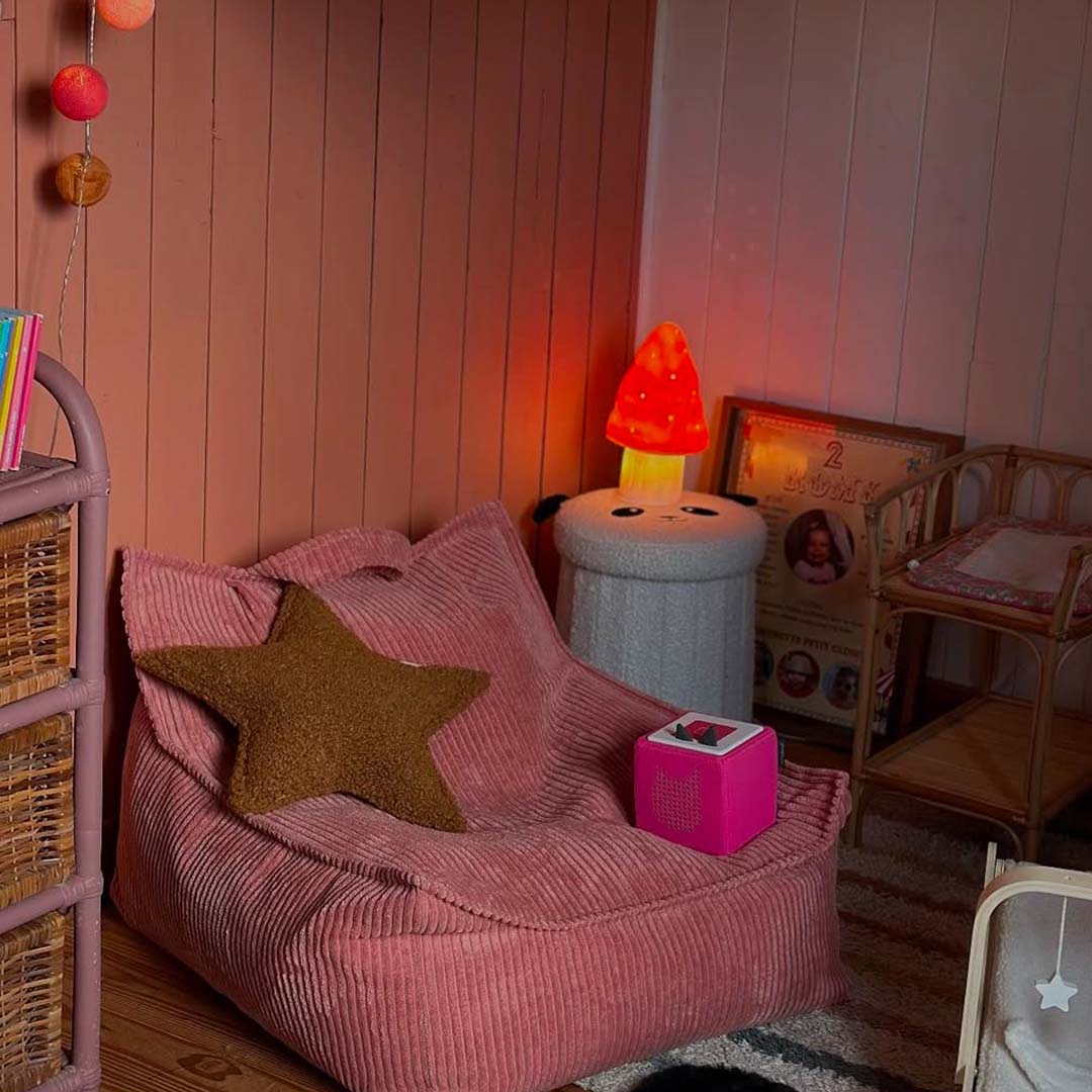Een verlichte paddenstoel nachtlamp op een krukje in een kinderkamer met een roze zitzak.