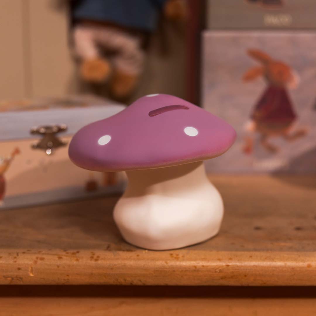 Lila paddenstoel spaarpot met witte stippen op een houten plank in een kinderkamer.