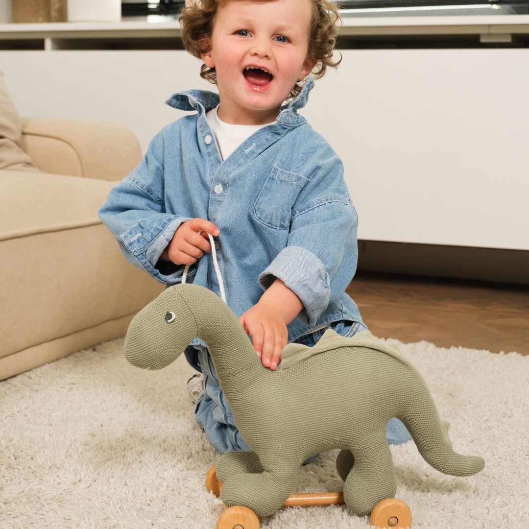 Een lachende peuter knielt op een vloerkleed en houdt de groene dinosaurus Victor trekfiguur vast.