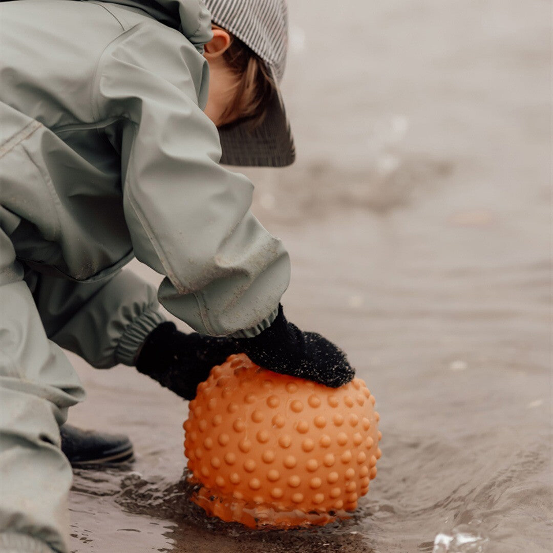 Een kind in een regenpak speelt met de oranje sensorische bal in ondiep water.