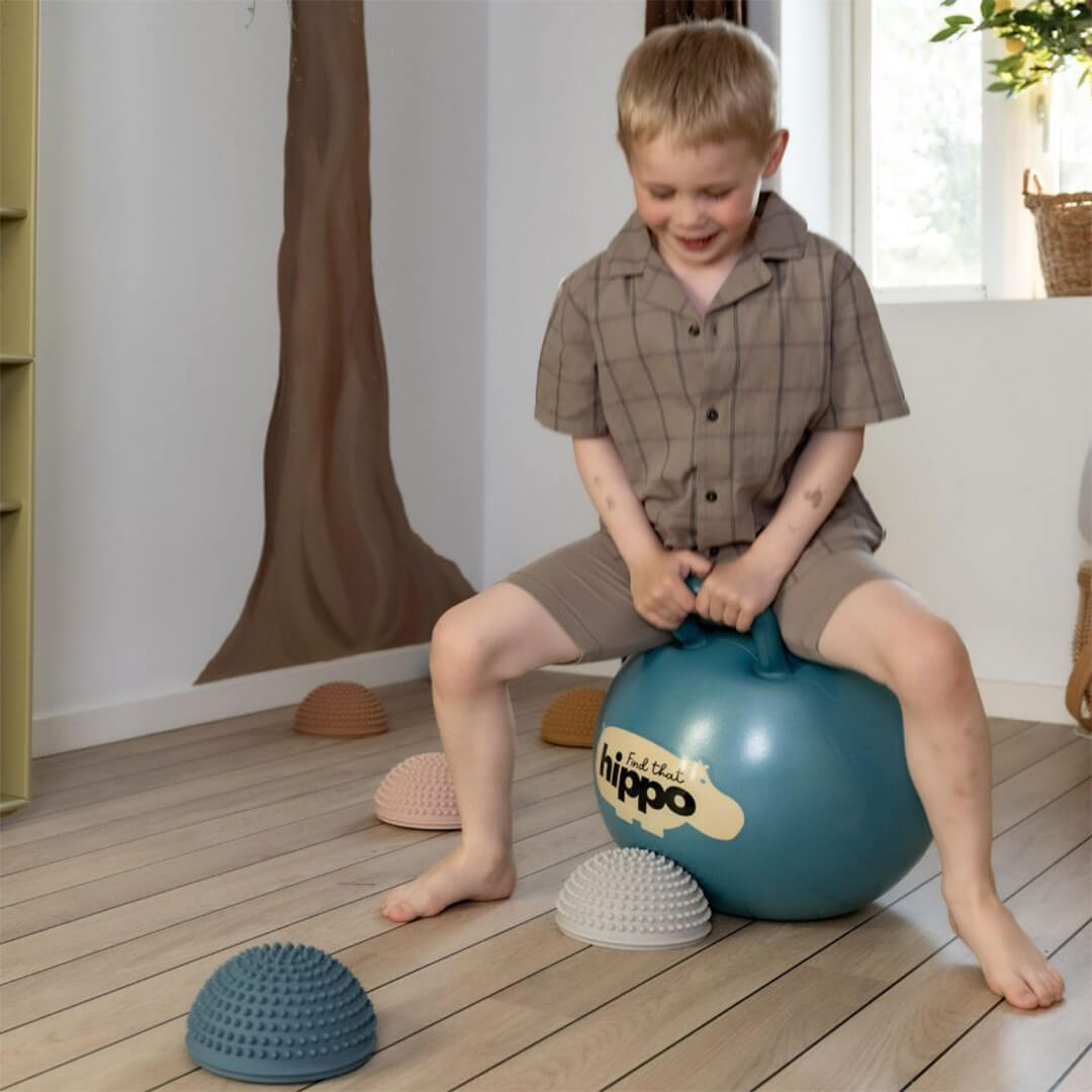 Een lachend kind zit op de find that hippo skippybal in een kamer met houten vloer.