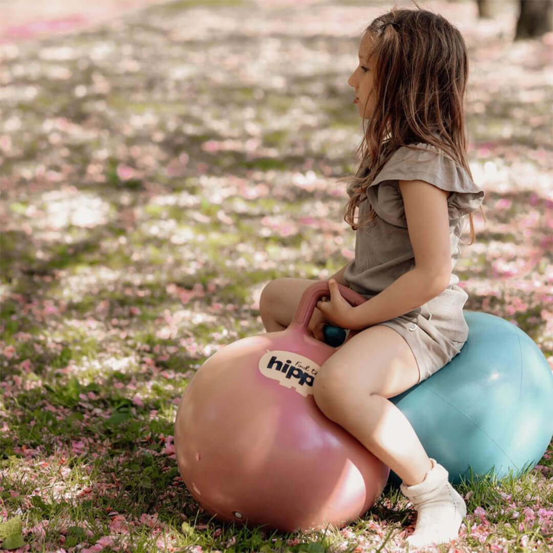Een meisje zit op een roze find that hippo skippybal in het gras met bloemblaadjes.