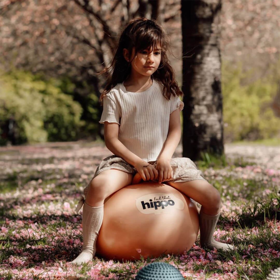 Een kind zit op de find that hippo skippybal in een park vol roze bloemblaadjes.