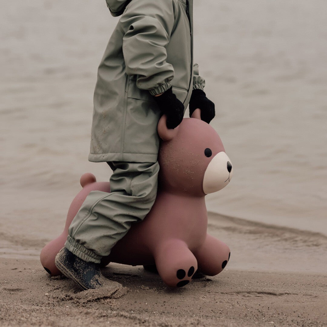Een kind in een regenpak staat op een zandstrand naast het find that hippo paarse skippydier beer.