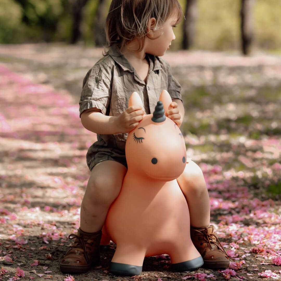 Een kind zit op een find that hippo roze eenhoorn skippydier in een park met roze bloemblaadjes op de grond.