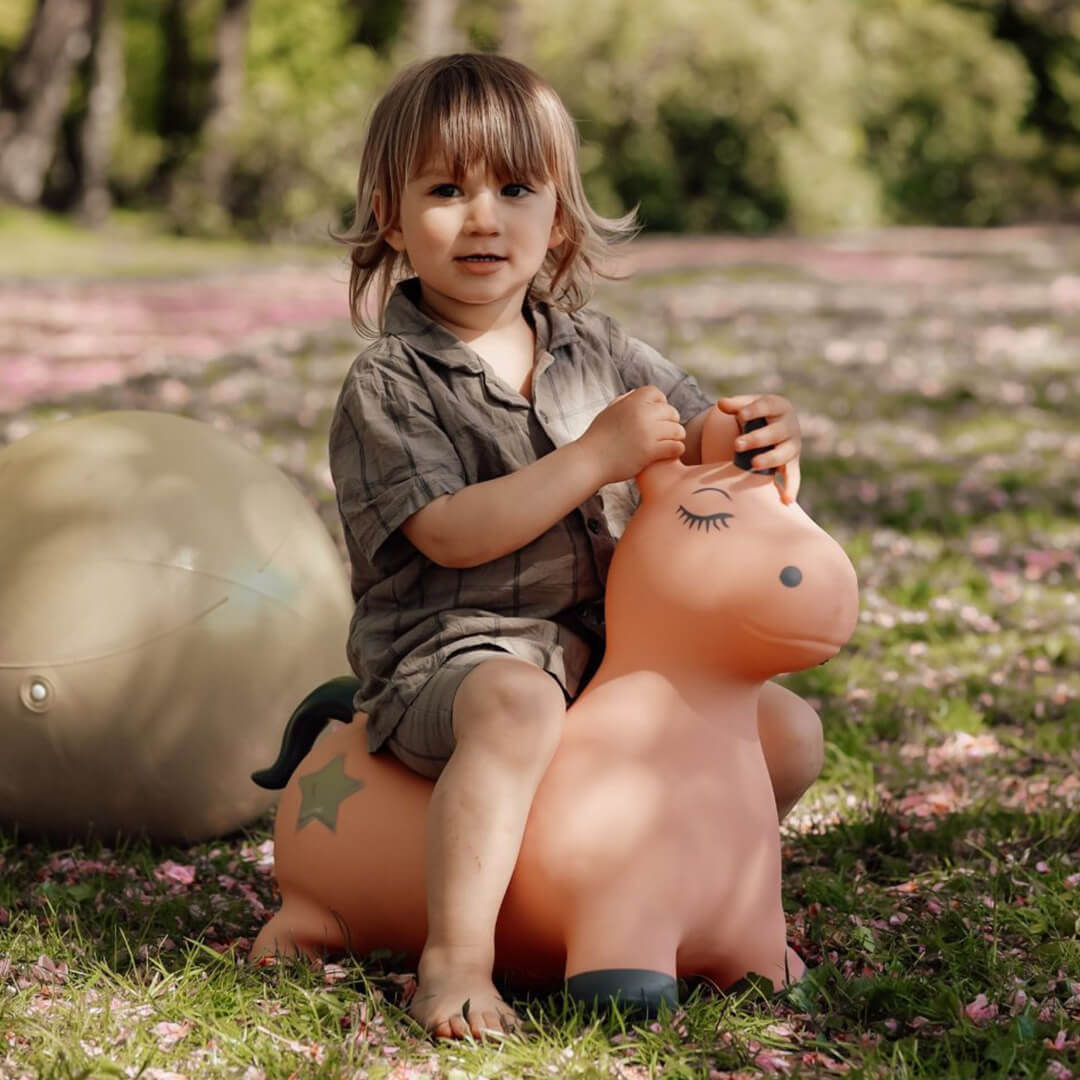 Een kind zit lachend op een find that hippo roze eenhoorn skippydier in het gras met roze bloemblaadjes.