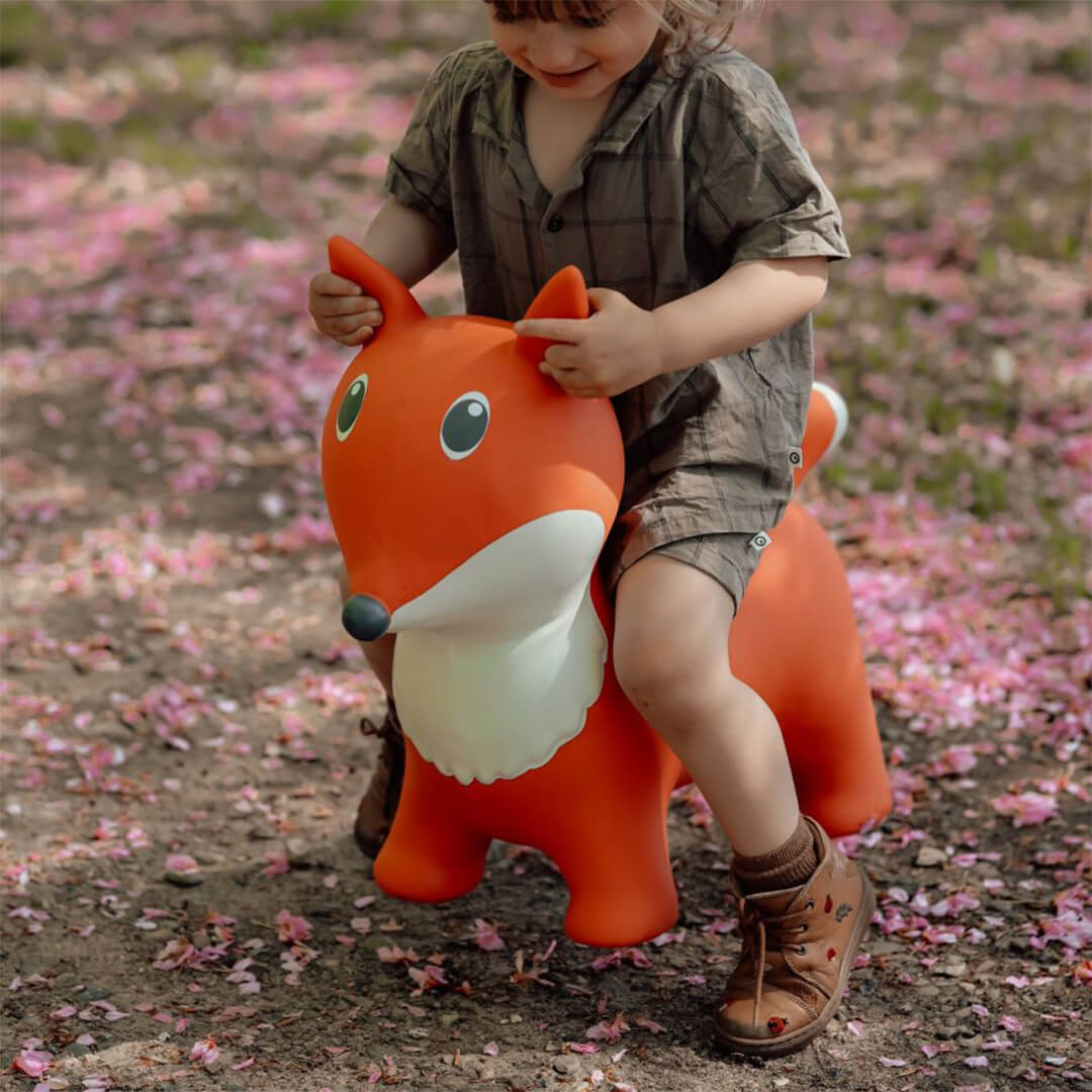 Een kind zit op de find that hippo skippydier vos, een oranje springkussen, buiten op een pad met roze bloemblaadjes.
