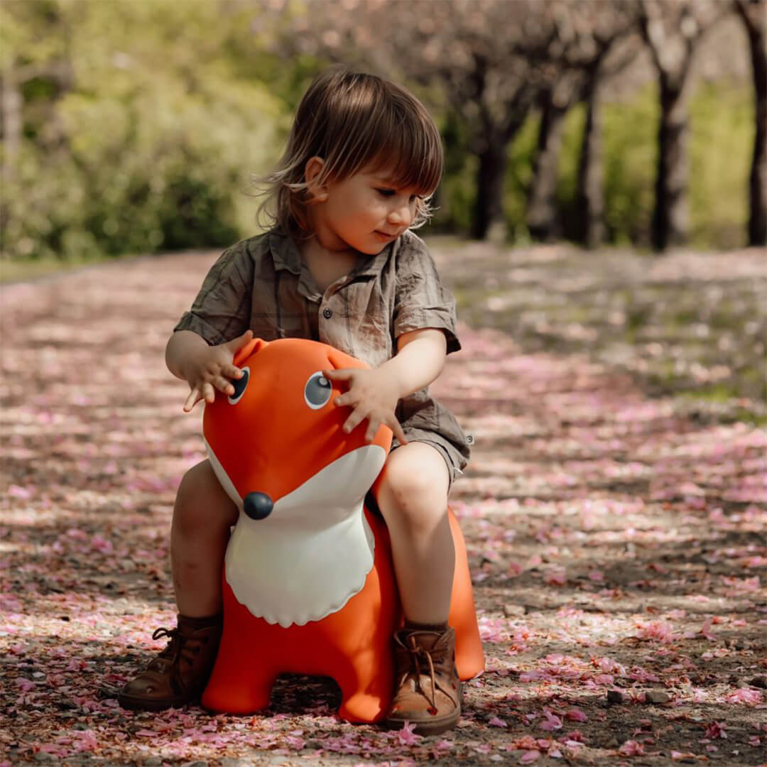 Een kind zit op de find that hippo skippydier vos, een oranje springkussen, op een pad met roze bloemblaadjes.