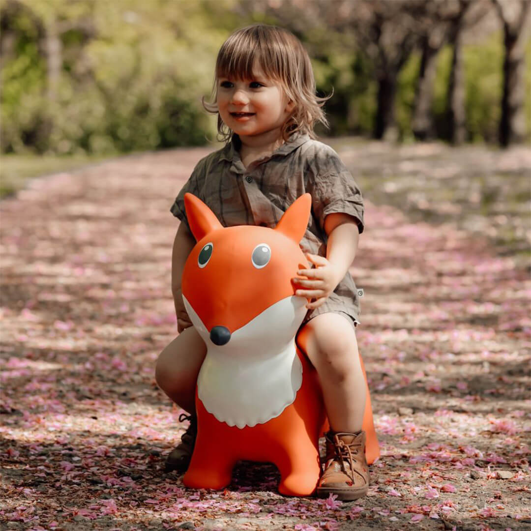 Een kind zit op de find that hippo skippydier vos, een oranje springkussen, van voren gezien op een pad met roze bloemblaadjes.