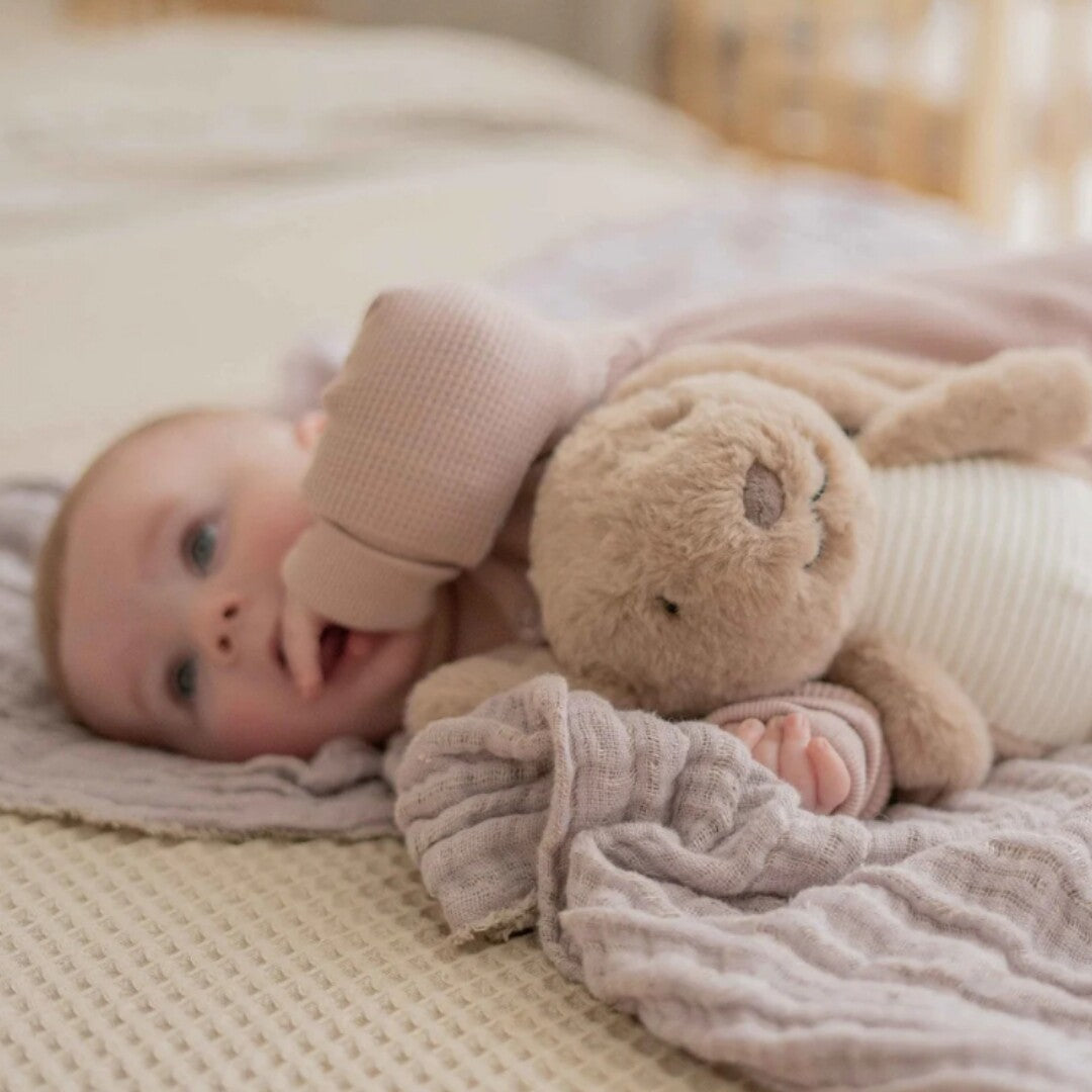 Een baby ligt op een deken met het bruine pluchen knuffelkonijn dichtbij haar arm.