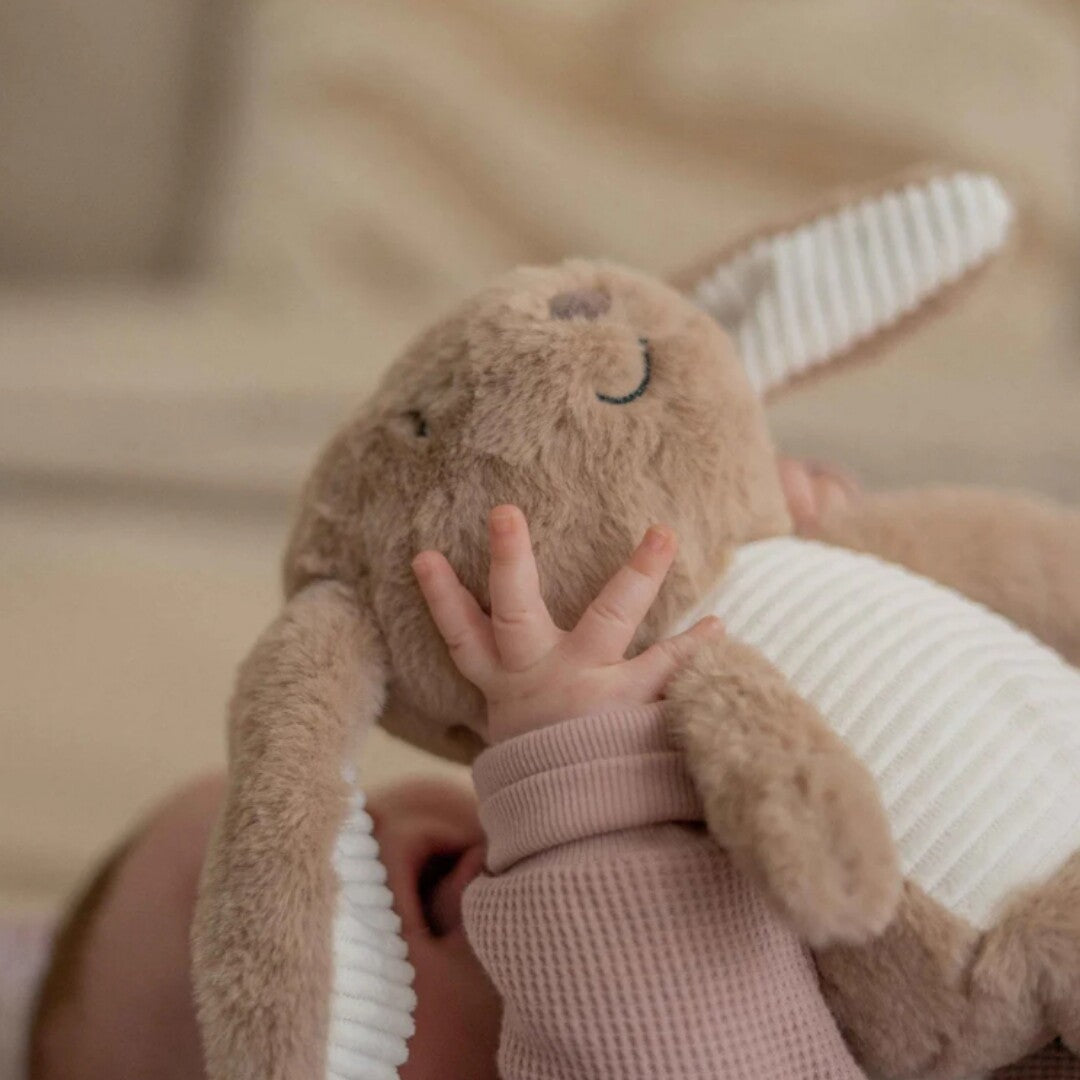 Een babyhandje pakt het bruine pluchen knuffelkonijn bij het hoofd.