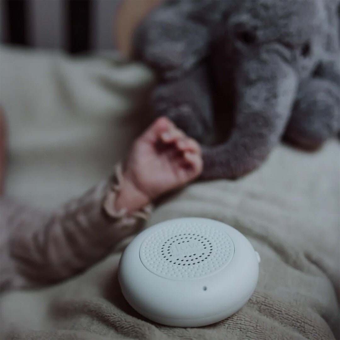 Witte hartslagmodule van flow amsterdam op een deken, met een babyhandje en knuffelolifant op de achtergrond.