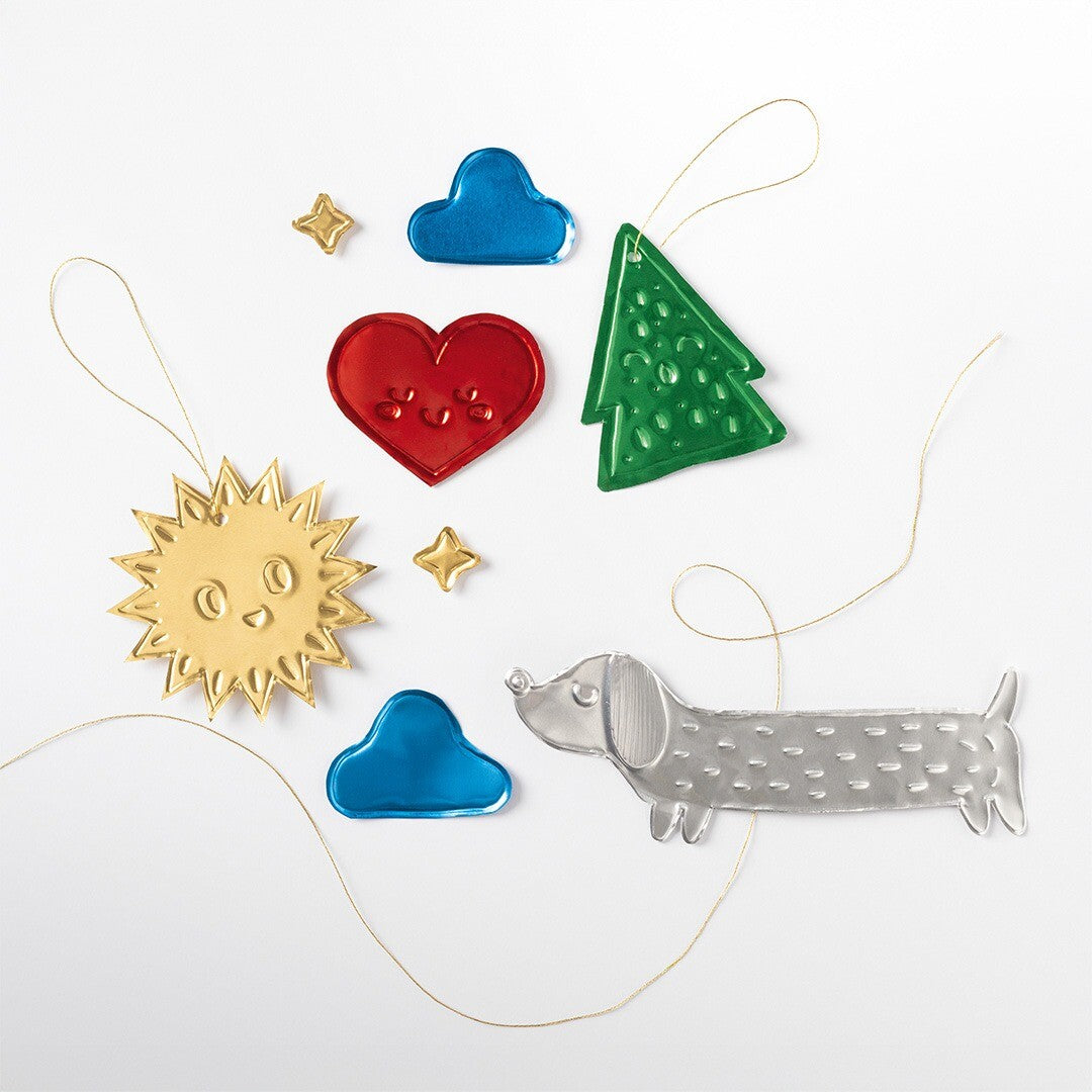 Een collectie van Folia reliëf aluminium figuren, waaronder een gouden zon, rood hart, blauwe wolken, groene kerstboom, zilveren hond en kleine sterren, op een witte achtergrond.
