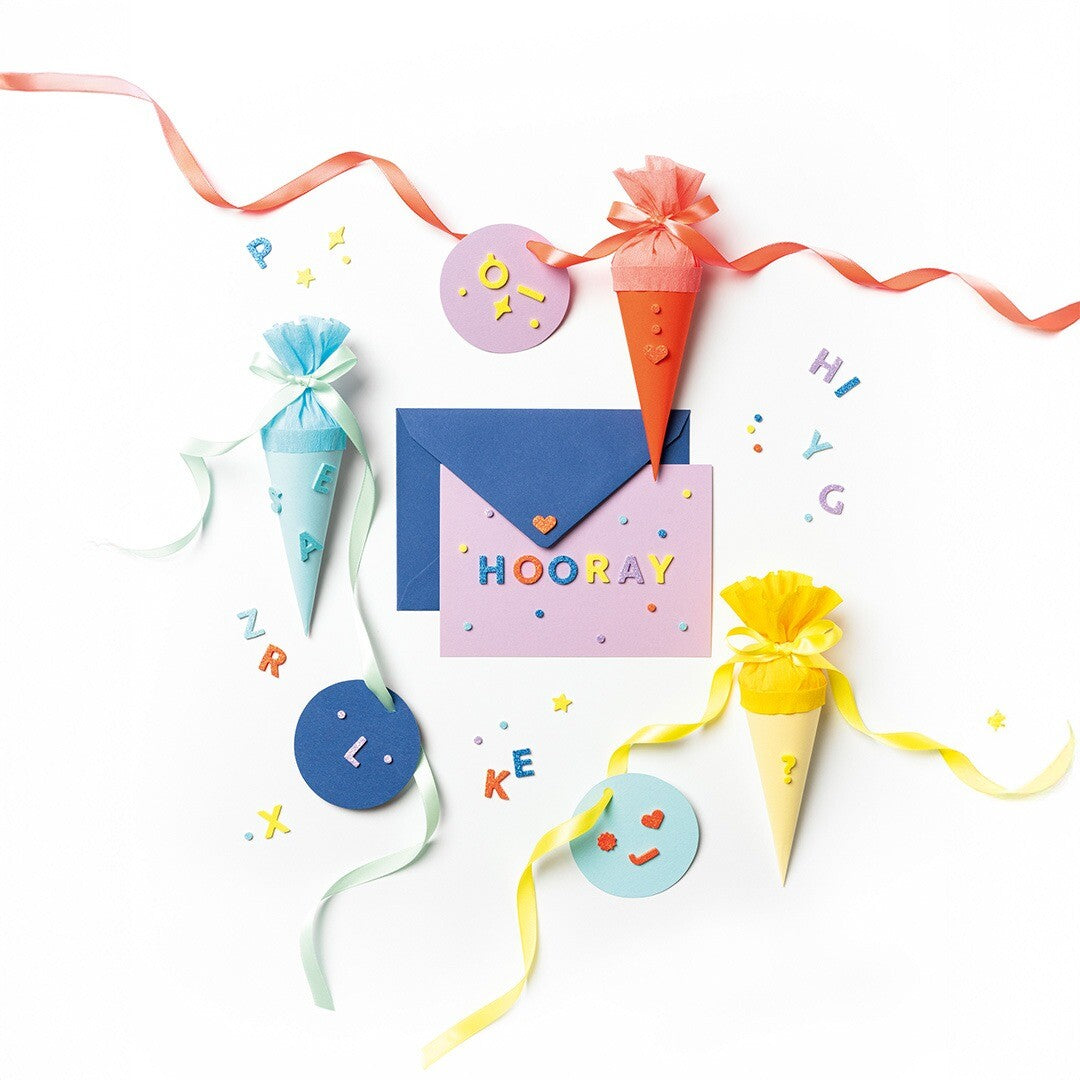 Creatieve demonstratie met Folia glitter letterstickers op een roze kaart met 'HOORAY', omringd door feestelijke puntzakjes en losse stickers.