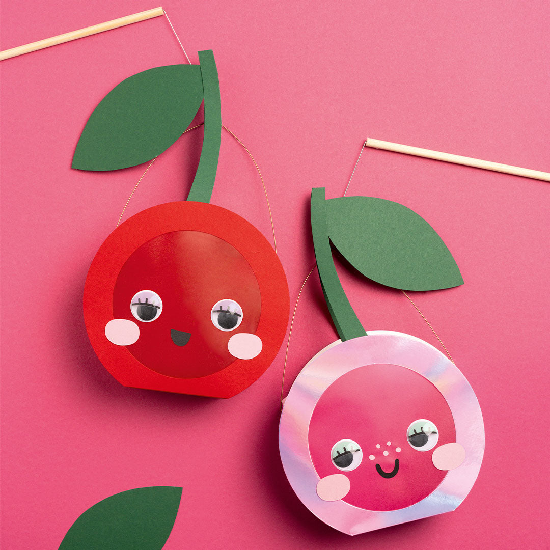 Twee knutselkersen met wiebelogen en wimpers, één rode en één roze, op een roze achtergrond.