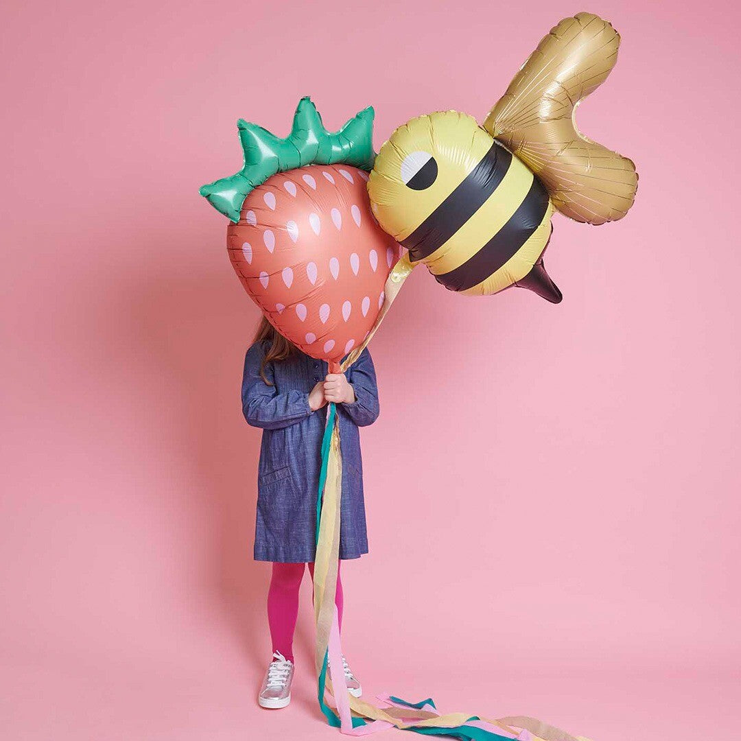 Een kind houdt een aardbei folieballon en een bij folieballon vast voor een roze achtergrond.