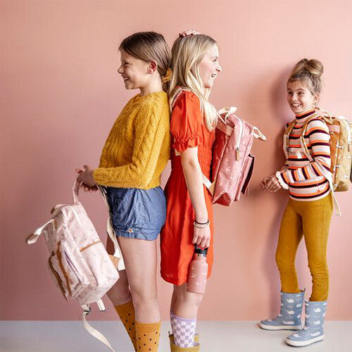 Drie lachende meisjes met rugzakken, staand tegen een roze muur.