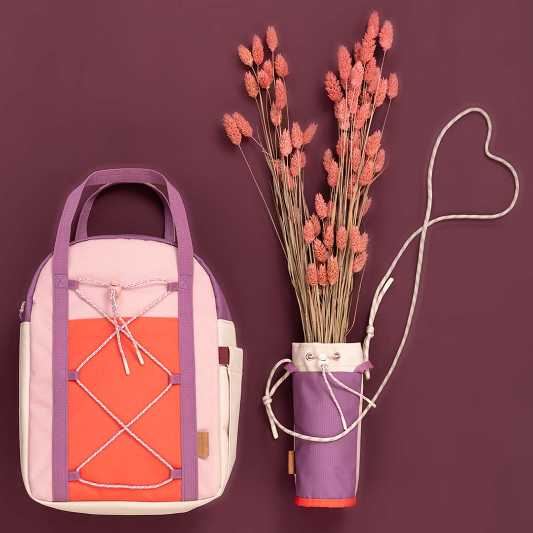 De Fresk peuterrugzak in roze, paars en oranje staat naast een bijpassende paarse waterfleshouder met gedroogde bloemen.