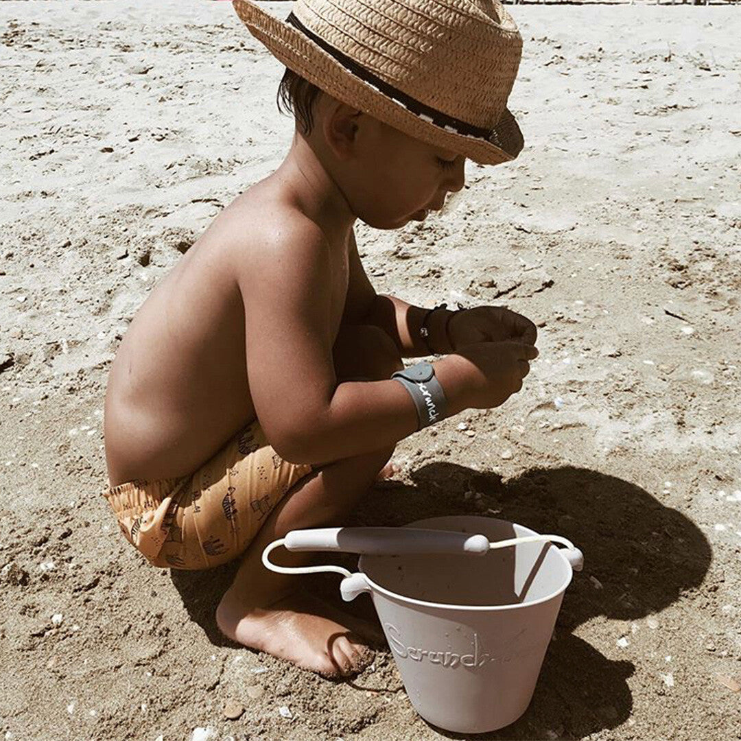 Een jongen met een strohoed speelt op het strand met een lichtgrijze Scrunch emmer en schep.