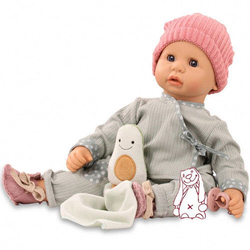 Götz babypop met roze muts, grijze kleding, speen en een avocado knuffel, zittend op de grond.
