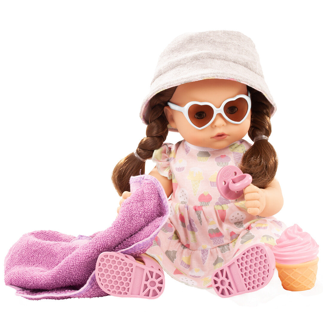 Götz babypop met bruine vlechten, roze jurk met ijsthema, grijze hoed, hartjeszonnebril, roze sandalen, handdoek en speen.