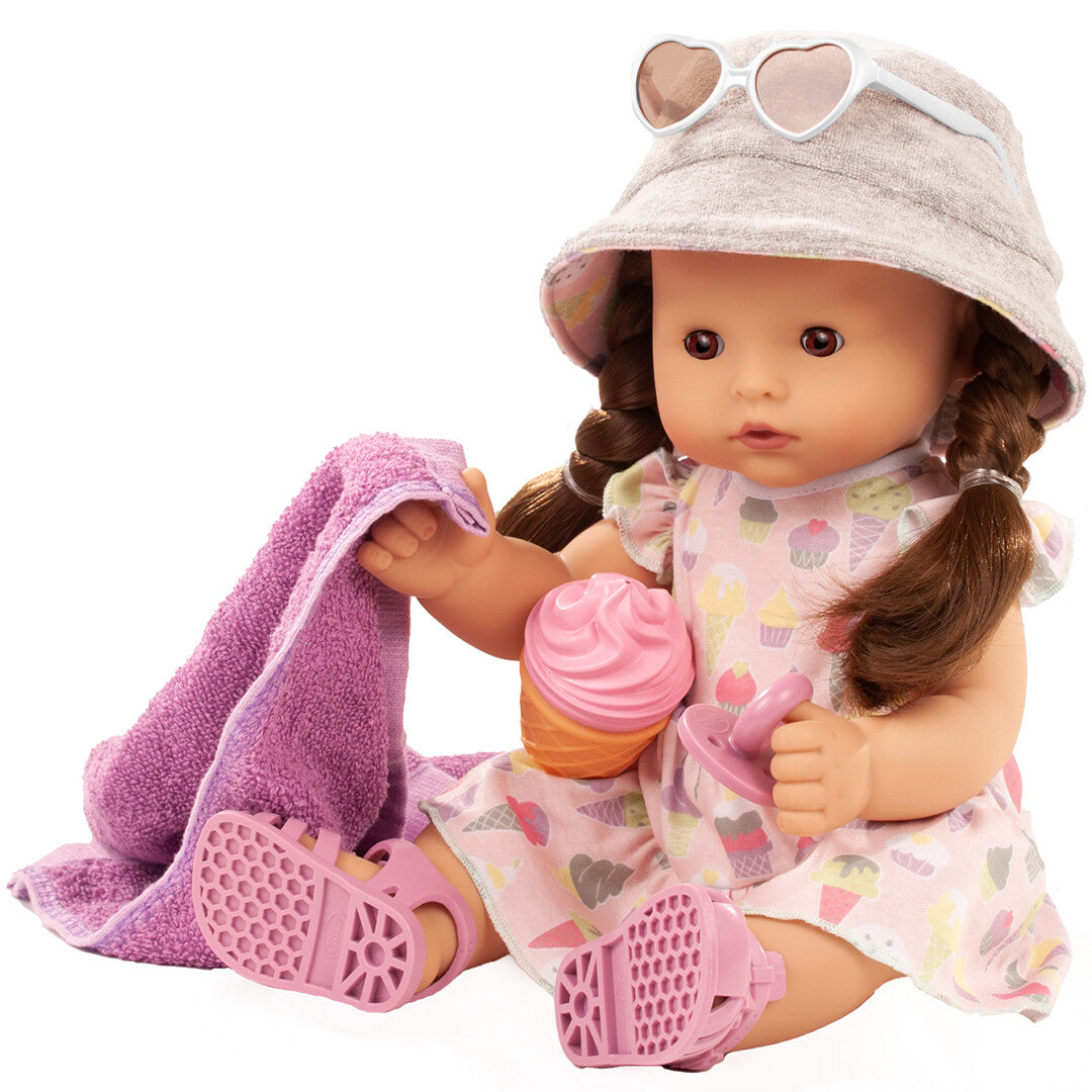 Götz babypop met bruine vlechten, roze jurk met ijsthema, grijze hoed, hartjeszonnebril op haar hoofd, roze sandalen, handdoek, speen en speelgoedijsje.