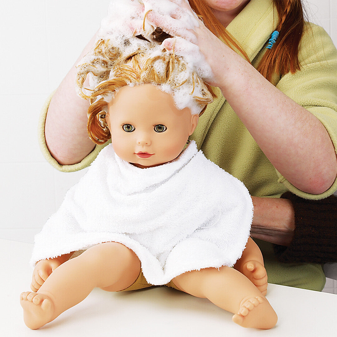Een babypop met blond haar wordt gewassen door een persoon, zittend op een witte ondergrond met een witte handdoek om.