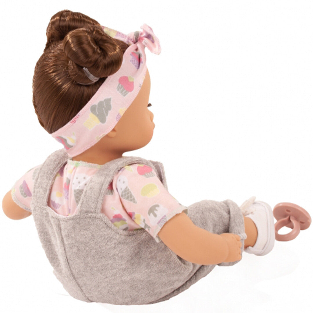 Achterkant van de babypop Muffin Popsicle zittend, met grijs tuinbroekje en roze shirt met cupcake-patroon.