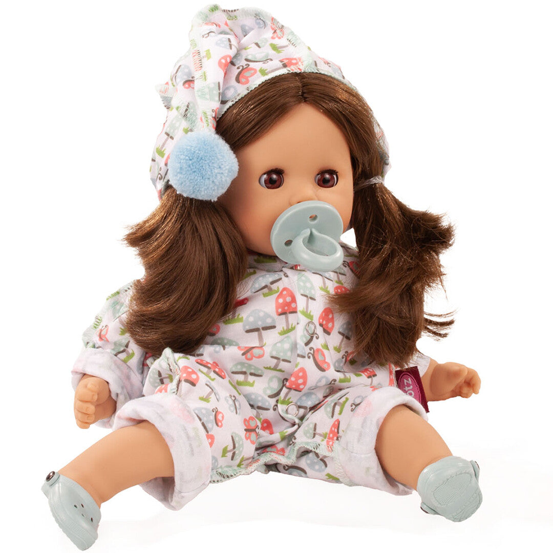 Gotz badpop met bruine pigtails, zittend in een wit slaappakje met paddenstoelenpatroon en bijpassende muts, met een blauwe speen in de mond.