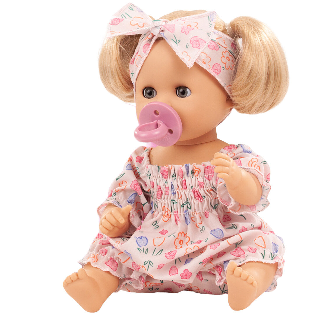 Götz badpop met blond haar, roze gebloemde outfit, hoofdband en roze speen, zittend schuin van voren.