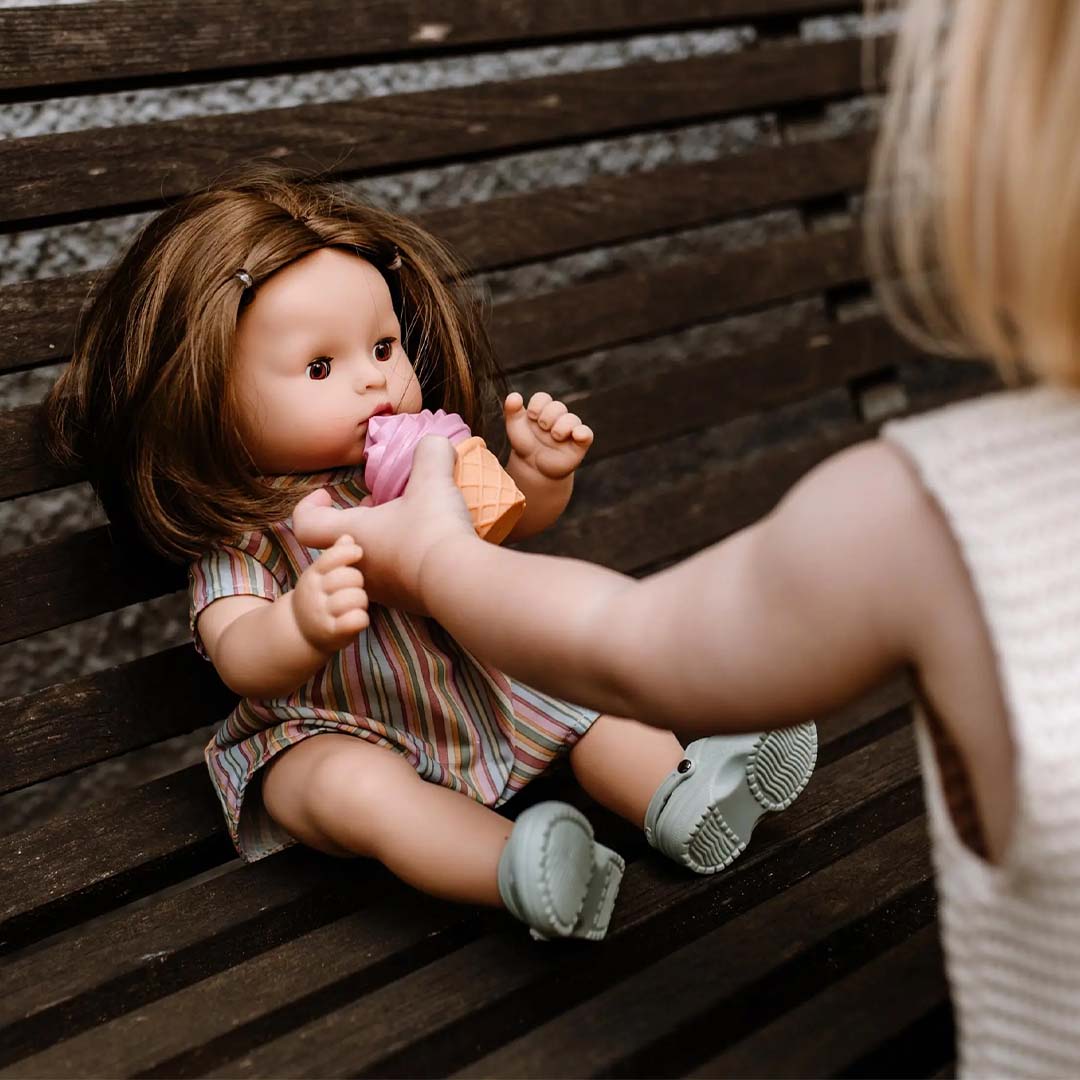 Een kind geeft de Götz babypop Maxy Aquini met haar een roze ijshoorntje terwijl ze op een houten bank zit.