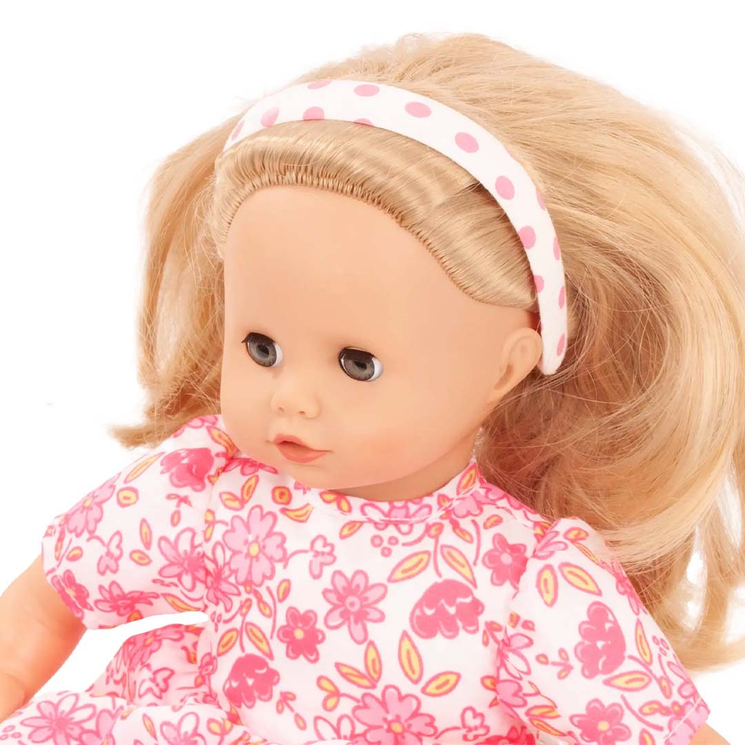 Een Gotz pop met blond haar draagt een witte haarband met roze stippen en een roze bloemetjesjurk.