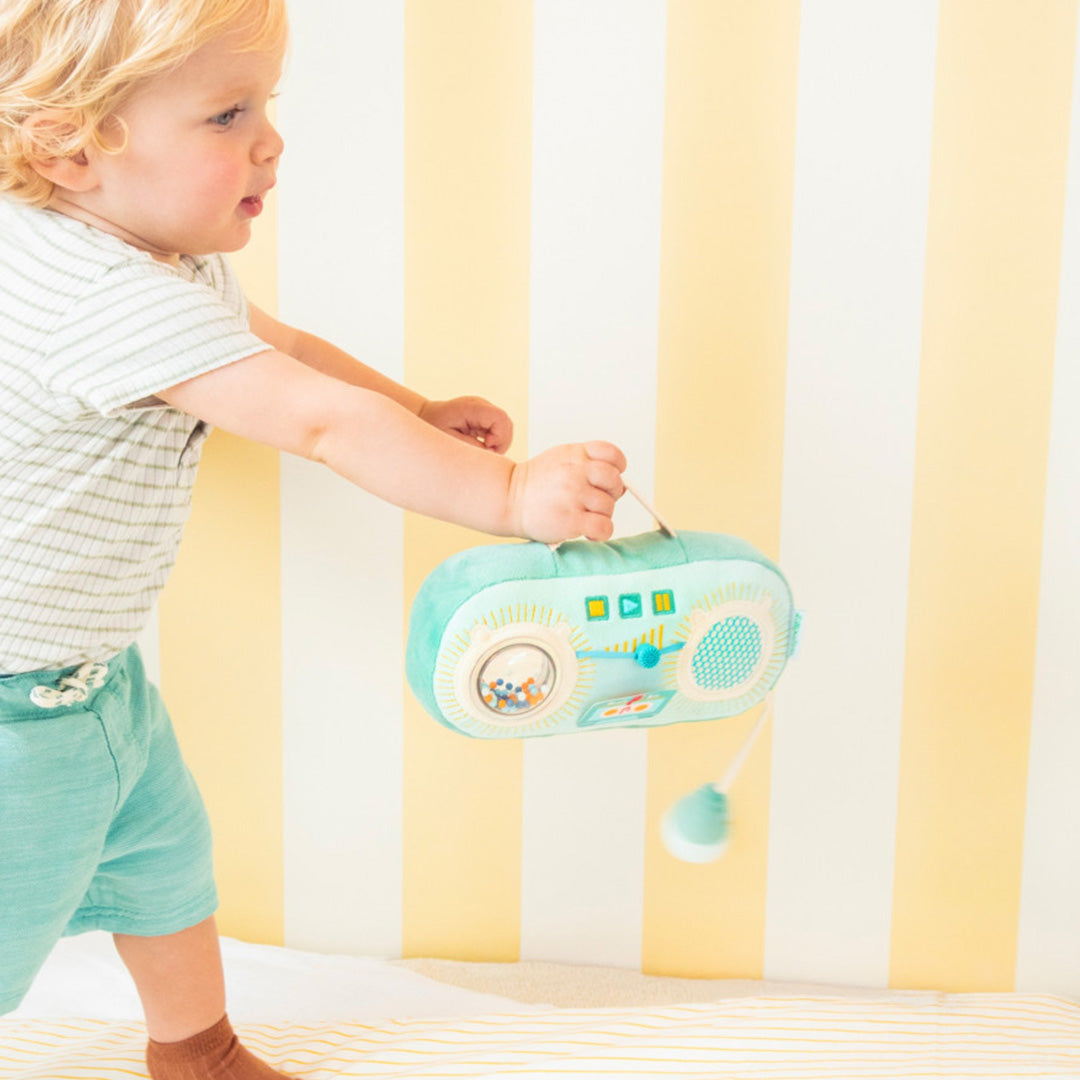 Een baby staat en draagt de Lilliputiens groene activiteitenradio aan het handvat.