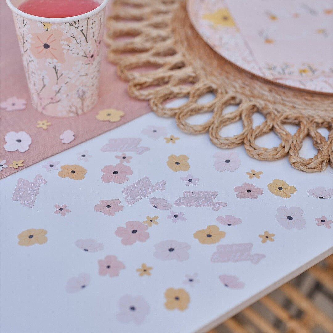 Ginger Ray confetti met bloemen en 'Happy Birthday' tekst verspreid over een tafel met bijpassende feestartikelen.