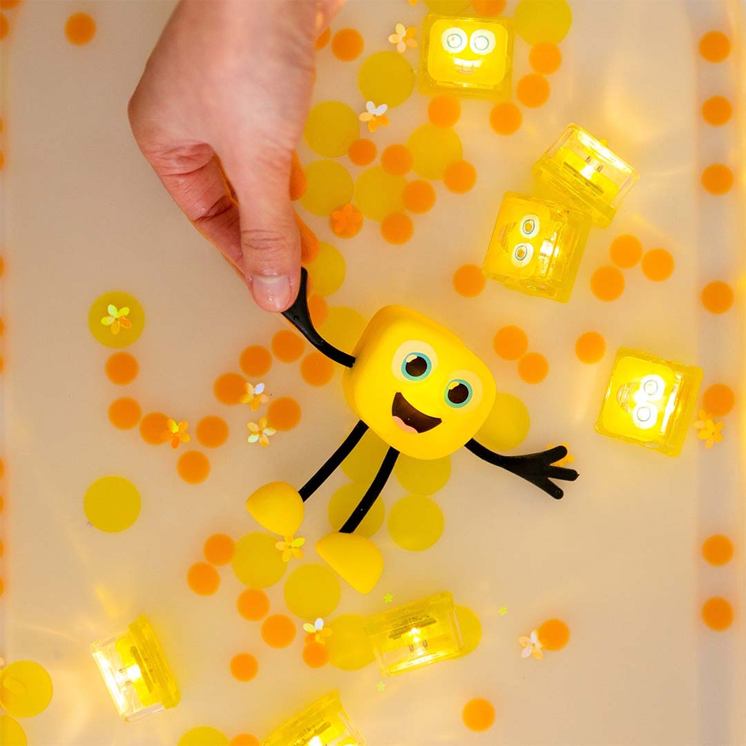 Een hand houdt het gele glo pals badspeelgoed Alex vast in een bad met lichtgevende kubussen en gele confetti.