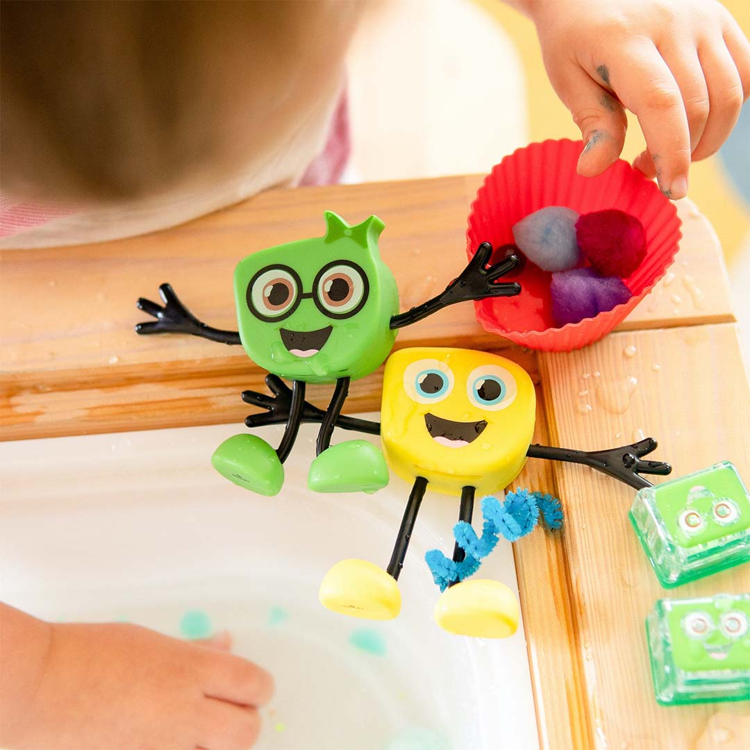 Twee glo pals badspeelgoedfiguren, een groene en de gele Alex, zitten bij een wasbak met lichtgevende kubussen.