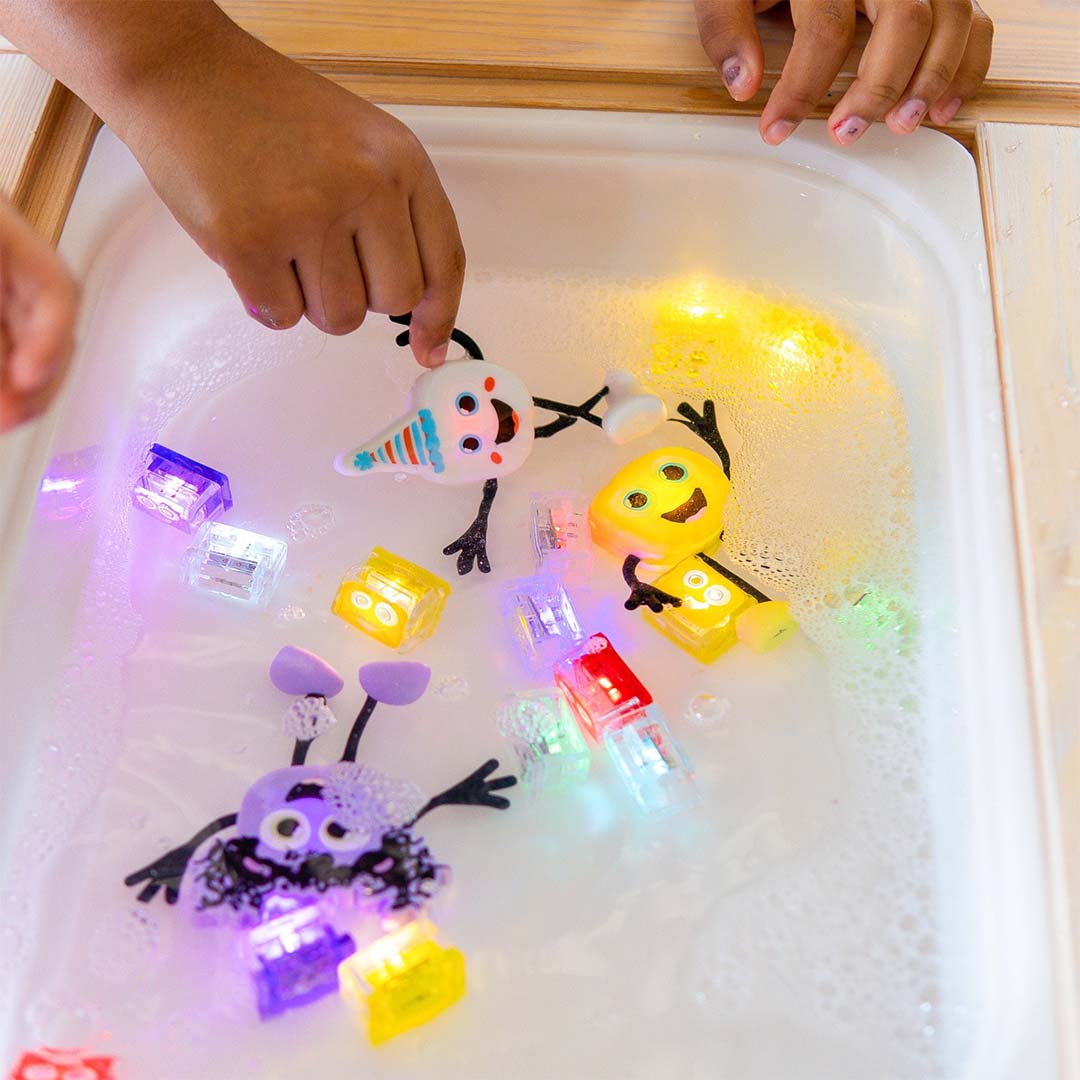 Kinderhanden spelen met verschillende Glo Pals lichtgevende badspeelgoedfiguren en lichtblokjes in schuimend water.