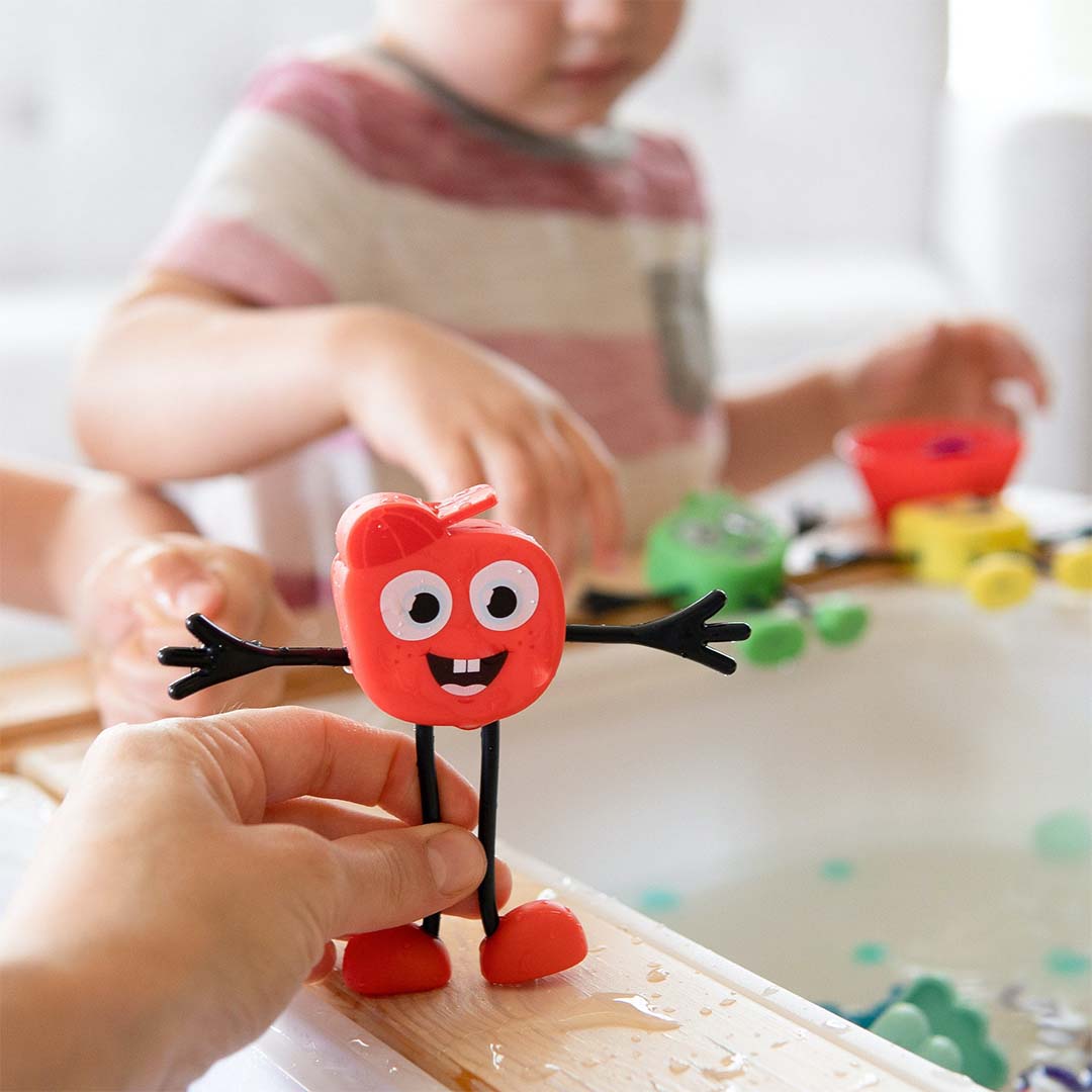 Een hand houdt het glo pals badspeelgoed Sammy vast, met een kind en andere speeltjes in een badkuip op de achtergrond.