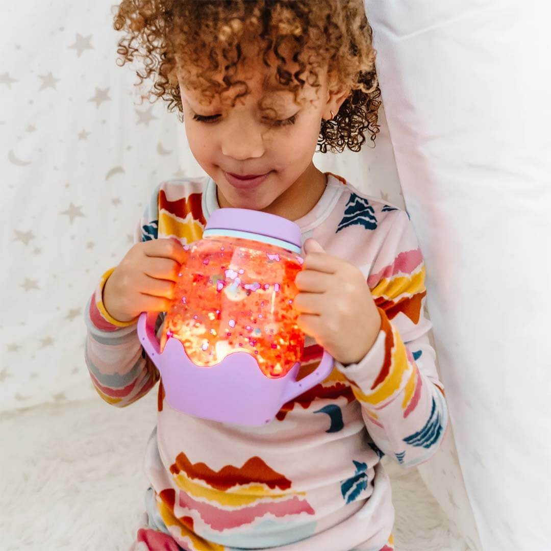 Een kind houdt een gloeiende paarse glo pals sensorische speelbeker met oranje glitter vast.