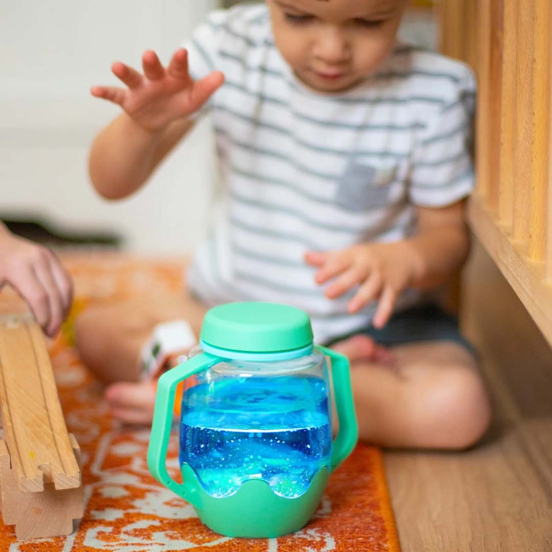 Een groene glo pals sensorische speelbeker met blauw gloeiend water staat op de grond.