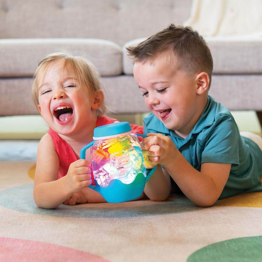 Twee lachende kinderen spelen samen met een gloeiende blauwe glo pals sensorische speelbeker op een kleurrijk kleed.