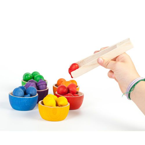 Een kinderhand gebruikt een houten tang om een rood houten eikeltje uit een rood bakje te pakken, onderdeel van het Grapat sorteerspeelgoed.