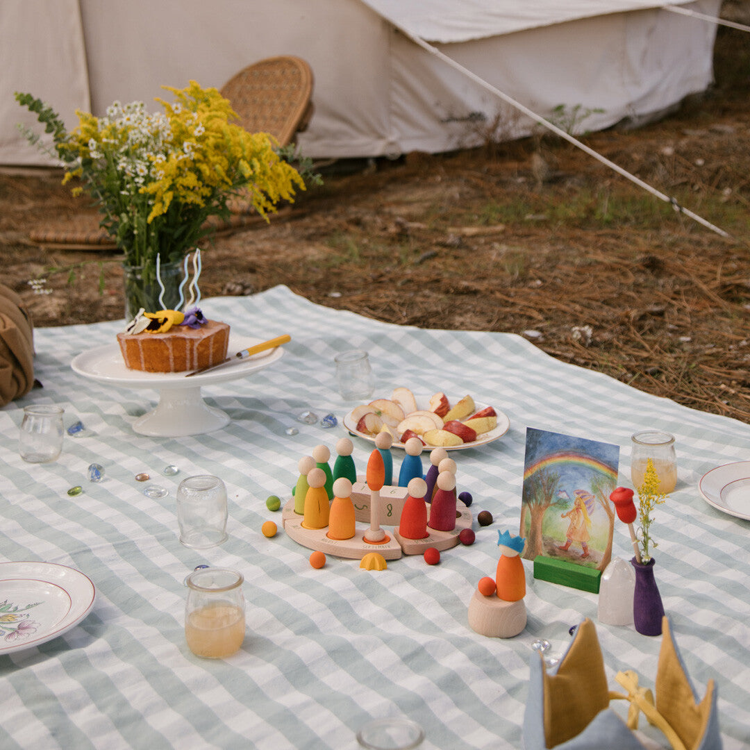Een picknicktafel met een Grapat eeuwige kalender, gekleurde poppetjes, een taart en fruit op een geruite deken.