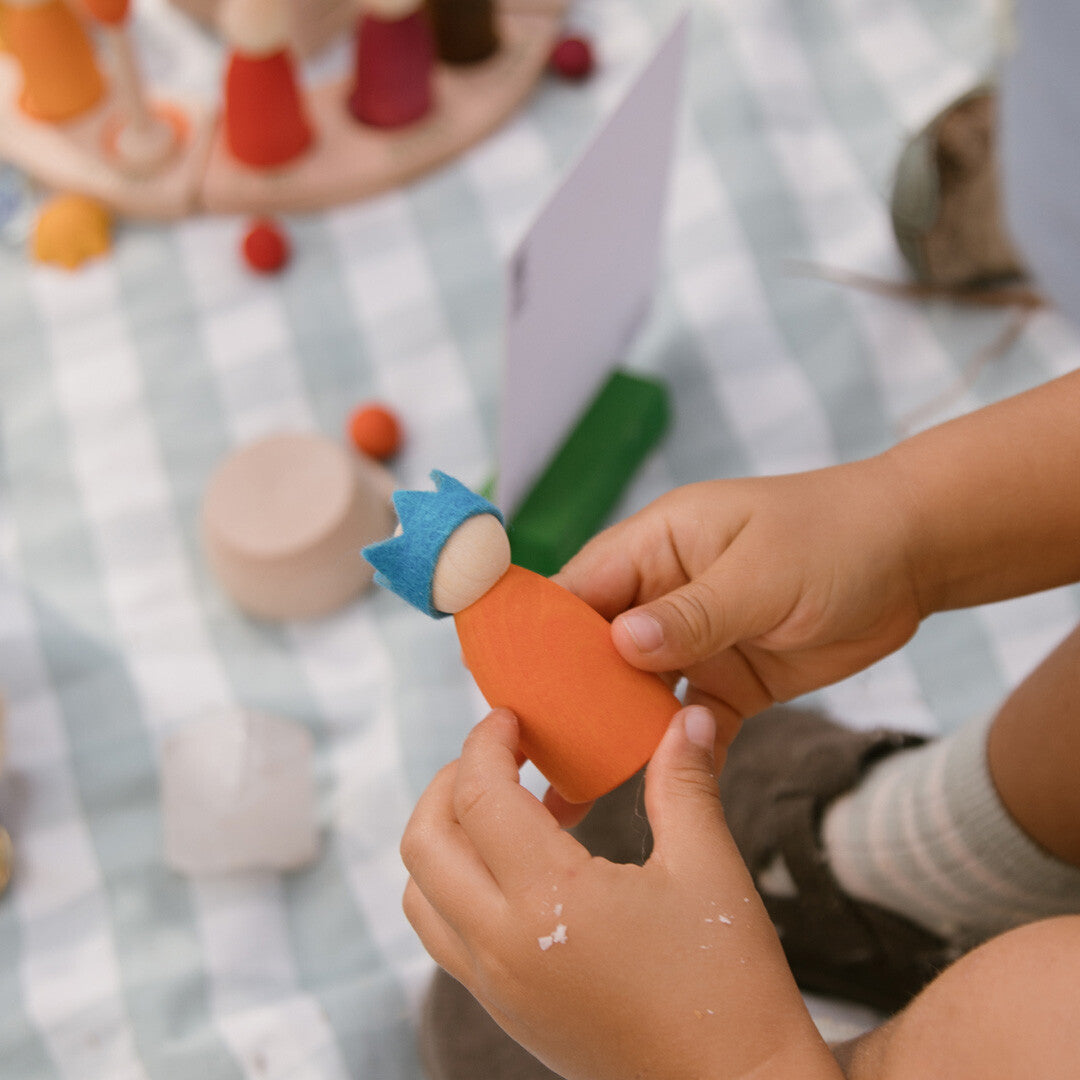 Kinderhanden houden een oranje houten poppetje met een blauwe vilten kroon vast.