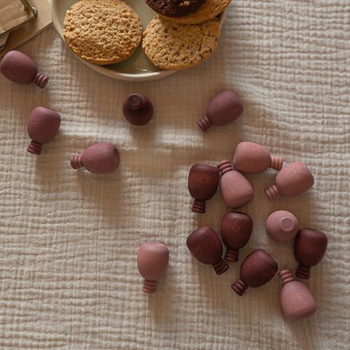 grapat houten speelfiguren in roze-paarse tinten verspreid over een lichte stof, met koekjes op de achtergrond.