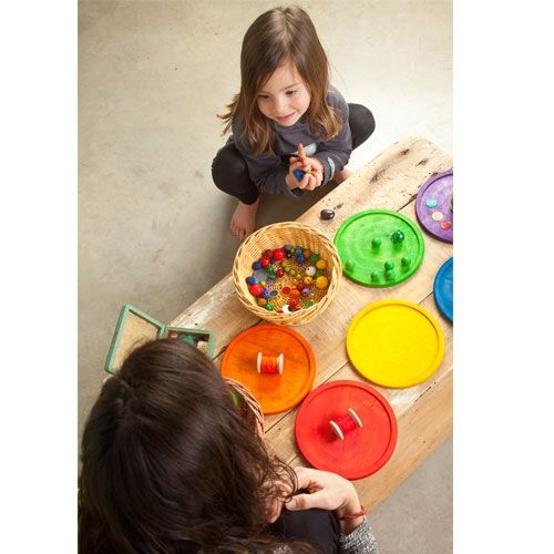 Een kind en een volwassene spelen met de kleurrijke houten sorteerplaten en kleine voorwerpen op een houten tafel.