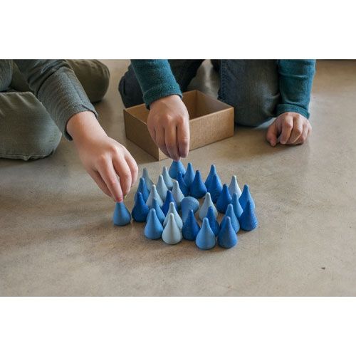Twee kinderhanden spelen met Grapat houten druppels in blauwtinten op de grond, met een kartonnen doos ernaast.