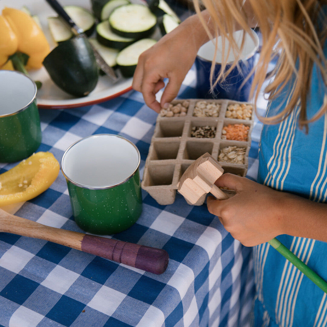 Een kind gebruikt een houten grapat stamper bij een tray met zaden op een blauw geruit tafelkleed.